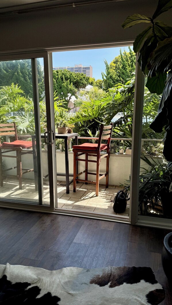 A room with a sliding glass door leading to an outdoor patio area. There is a wooden chair with a red cushion, a table, and a potted plant.
