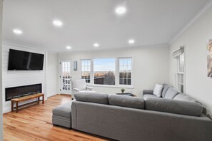 A modern living room with a grey sectional sofa, a flat-screen TV mounted on the wall, and a wooden bench. - Hilltop Retreat (Round Hill)