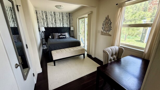 A bedroom with a dark-colored bed, a white bench, a wooden table, and a window with a view of greenery. - Discover the charm of Palmer Lake at this cozy cottage in this peaceful town. (Palmer Lake)