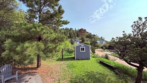 A small shed in a grassy area with trees and a bench nearby. - Discover the charm of Palmer Lake at this cozy cottage in this peaceful town. (Palmer Lake)