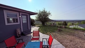 A patio with two red chairs and a blue table, a gray door, and a view of a rural landscape. - Discover the charm of Palmer Lake at this cozy cottage in this peaceful town. (Palmer Lake)