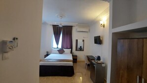 A hotel room with a bed, a desk, a chair, an air conditioning unit, and a television.