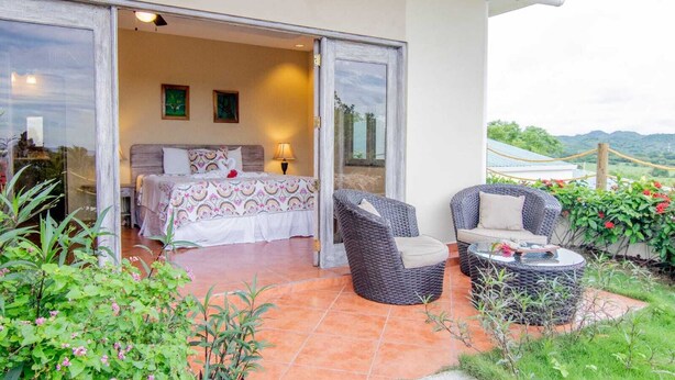 A bedroom with a bed, a nightstand, and a lamp. There is a balcony with wicker furniture and a view of the landscape. - king Standart pool view (Tola)