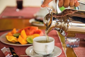 Une main verse une boisson d’un pichet métallique dans une tasse blanche posée sur une soucoupe.