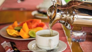 A hand pouring a beverage from a metallic pitcher into a white cup on a saucer. - Serenity Boutique Hotel (Paje)