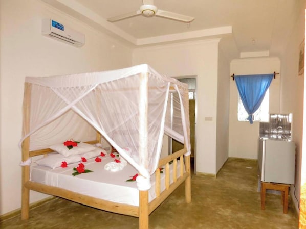 Une chambre individuelle avec un lit en bois, du linge de lit blanc, un ventilateur de plafond, un appareil de climatisation et une petite kitchenette.