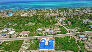 An aerial view of a coastal residential area with houses, a road, and a large blue-roofed building. - Serenity Boutique Hotel (Paje)