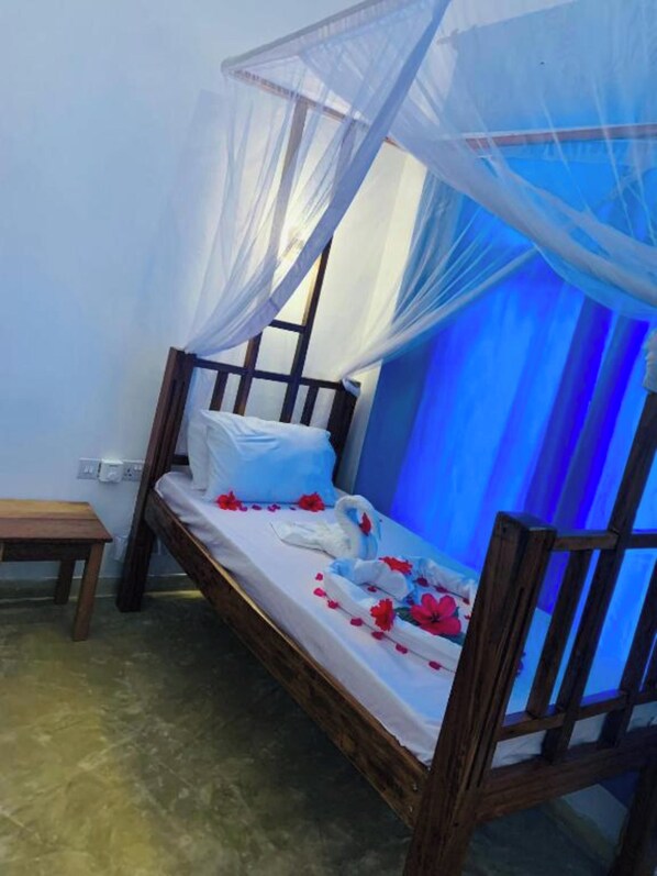 A single bed with a wooden frame and a mosquito net, a small wooden table, and a wall socket.