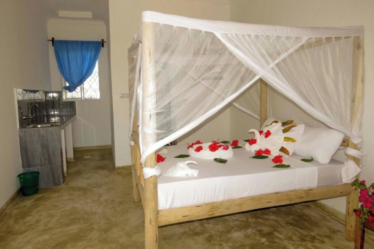 A single bed with a white canopy, a small table with a vase of flowers, and a kitchenette in the background.
