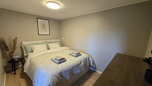 Ein ordentlich bezogenes Bett mit einem gemusterten Überwurf und Kissen, ein gerahmtes Kunstwerk an der Wand und ein kleiner Nachttisch mit einer Vase getrockneter Pflanzen.