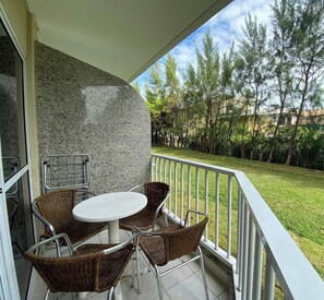 A balcony with a white table and wicker chairs, a stone wall, and a view of a grassy area with trees.