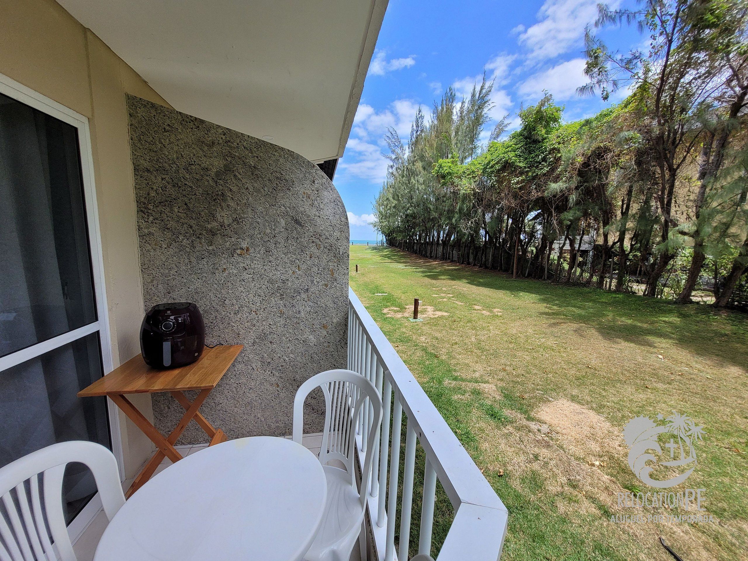 A balcony with a table and chairs, a speaker, and a view of a grassy area with trees.