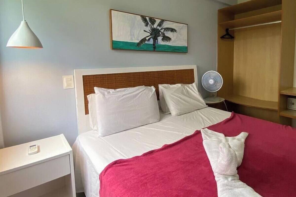 A hotel room with a bed, a bedside table, a wall-mounted fan, and a painting of a palm tree.