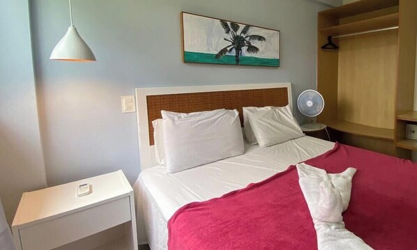 Ein Hotelzimmer mit Bett, Nachttisch, Wandventilator und einem Palmenbaumgemälde.