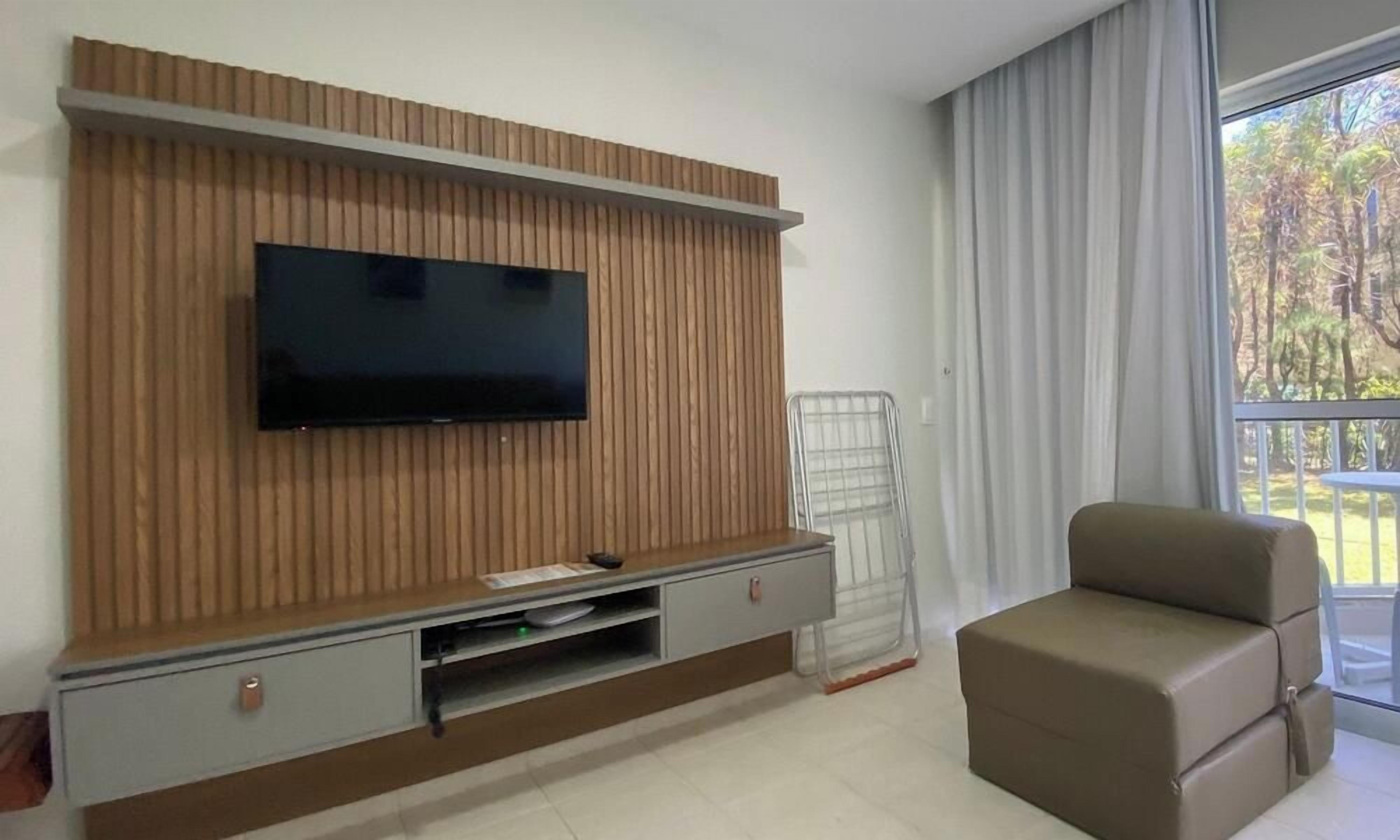 A modern living room with a flat-screen TV mounted on a wooden panel wall, a grey sofa, and a glass door leading to an outdoor area.