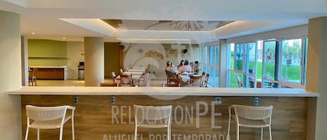 Un área de recepción moderna con un mostrador de madera, taburetes de mimbre blanco y vistas al exterior.