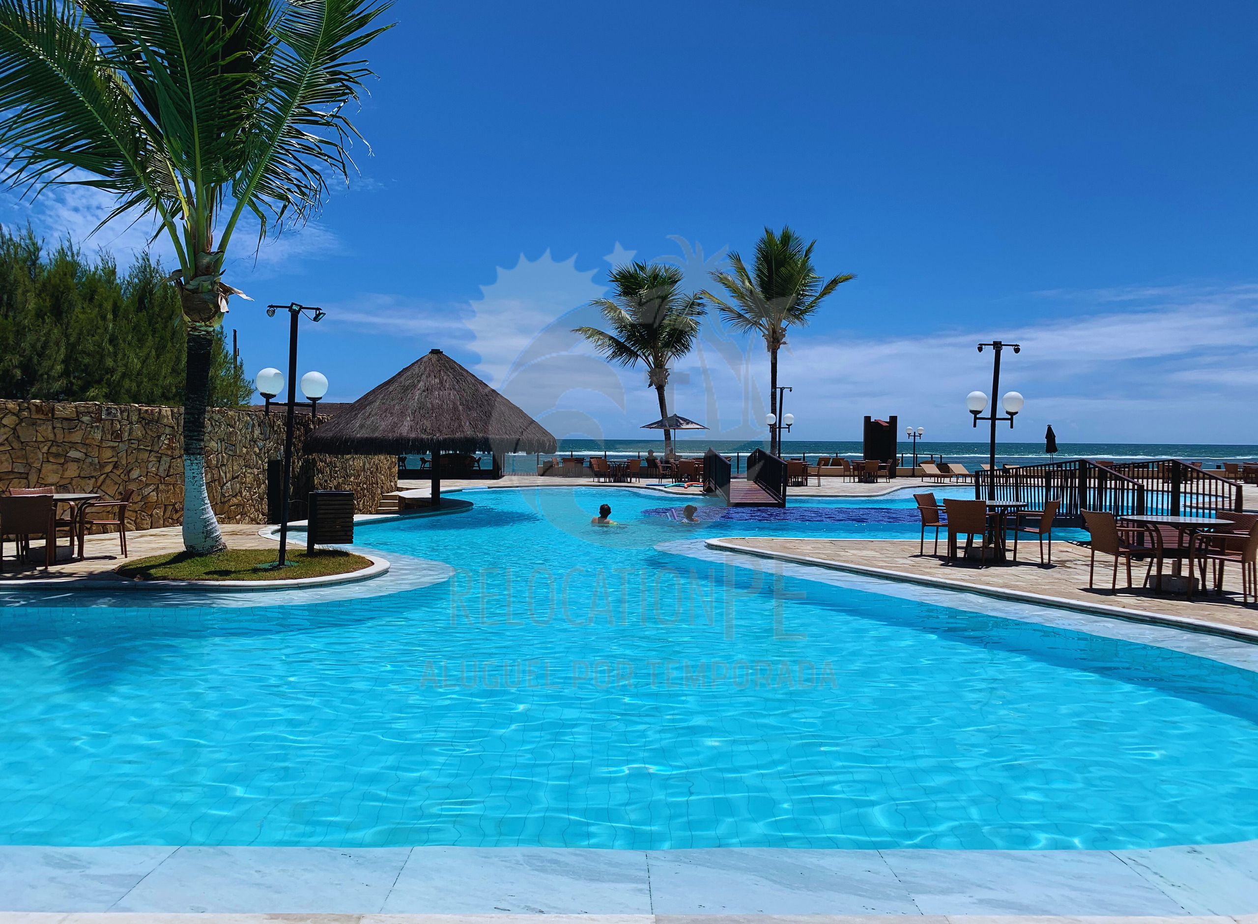 A resort pool with a thatched-roof structure, palm trees, and a view of the ocean.