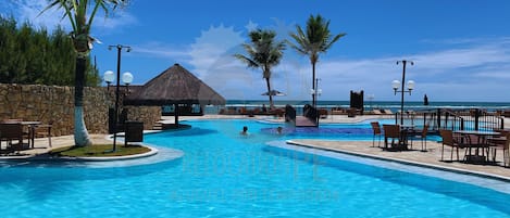 A resort pool with a thatched-roof structure, palm trees, and a view of the ocean.