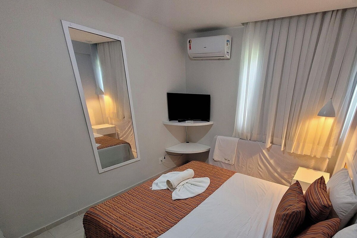 A hotel room with a bed, a TV, a mirror, a bedside table, a window with curtains, and a wall-mounted air conditioner.