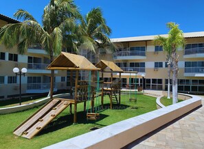 A playground with slides and climbing structures, surrounded by palm trees and a multi-story building with balconies. - Marupiara Suites (Ipojuca)