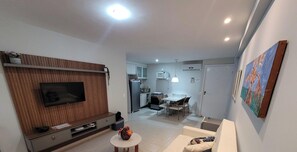 A modern living room with a flat-screen TV, a dining area with a table and chairs, and a kitchen in the background. - Marupiara Suites (Ipojuca)