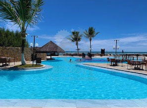 A resort pool with a thatched-roof structure, palm trees, and a view of the ocean.