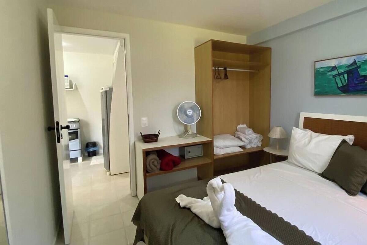 A hotel room with a bed, a fan, a shelf with towels, a painting, and a door leading to another room.