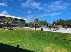 A resort with a swimming pool, outdoor seating, a playground, and a building with balconies.