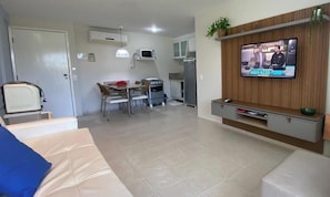 A modern living room with a sofa, a dining table with chairs, a kitchen area with a microwave, oven, and refrigerator, and a TV mounted on the wall.