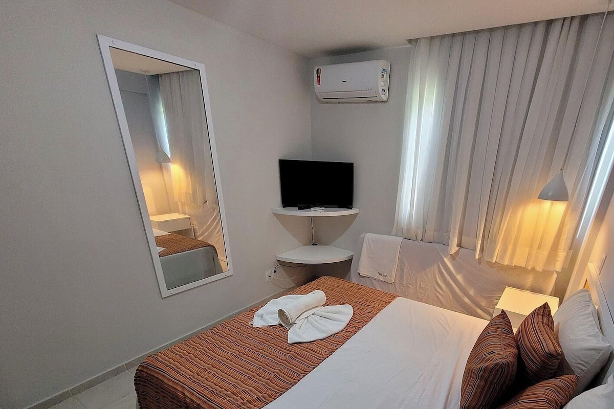 Habitación de hotel con cama, televisor, espejo, lavabo y ventana con cortinas.