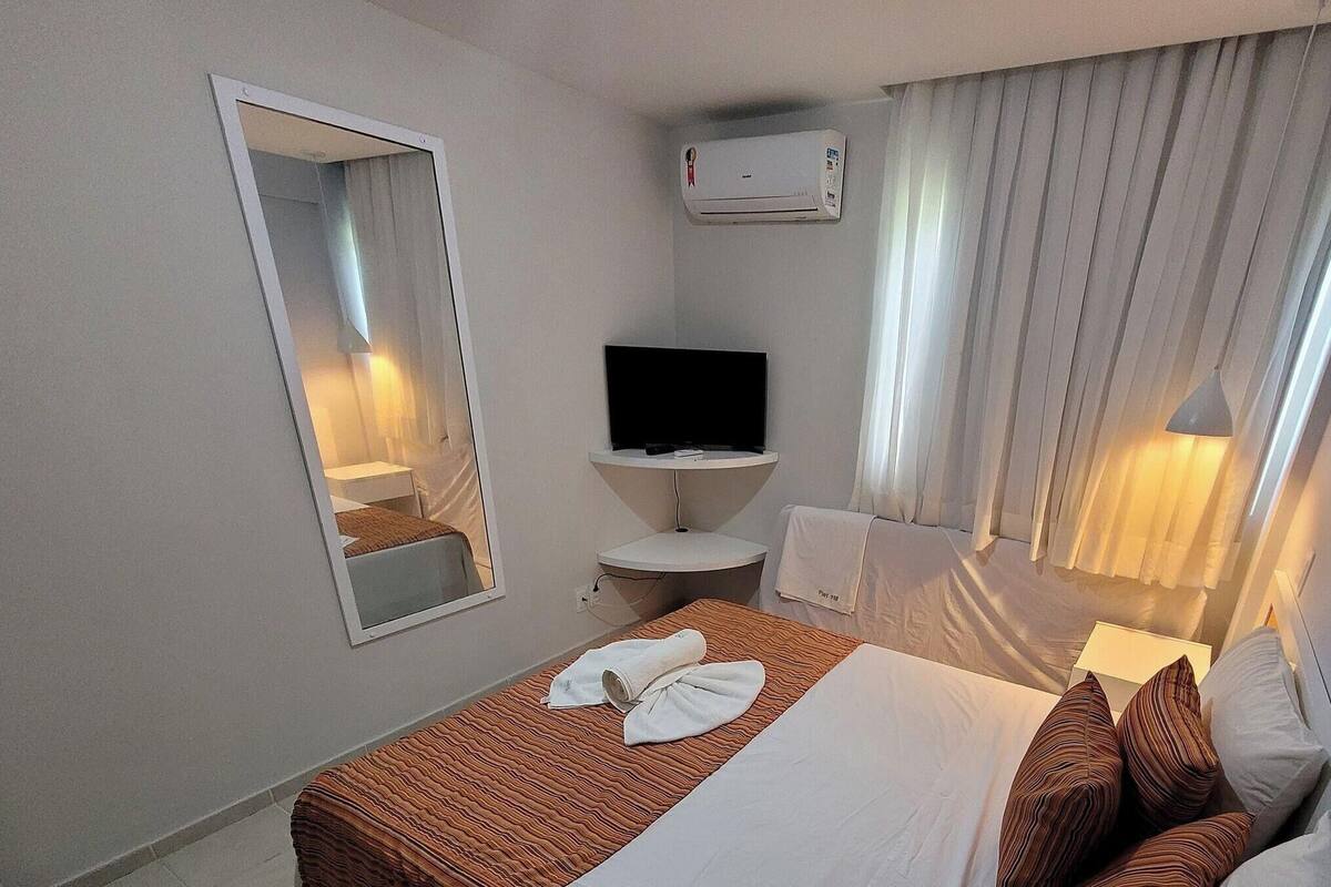 A hotel room with a bed, a TV, a mirror, an air conditioner, a sink, and a window with curtains.