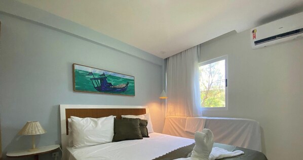 A hotel room with a bed, a painting of a boat, a window with curtains, and an air conditioning unit.