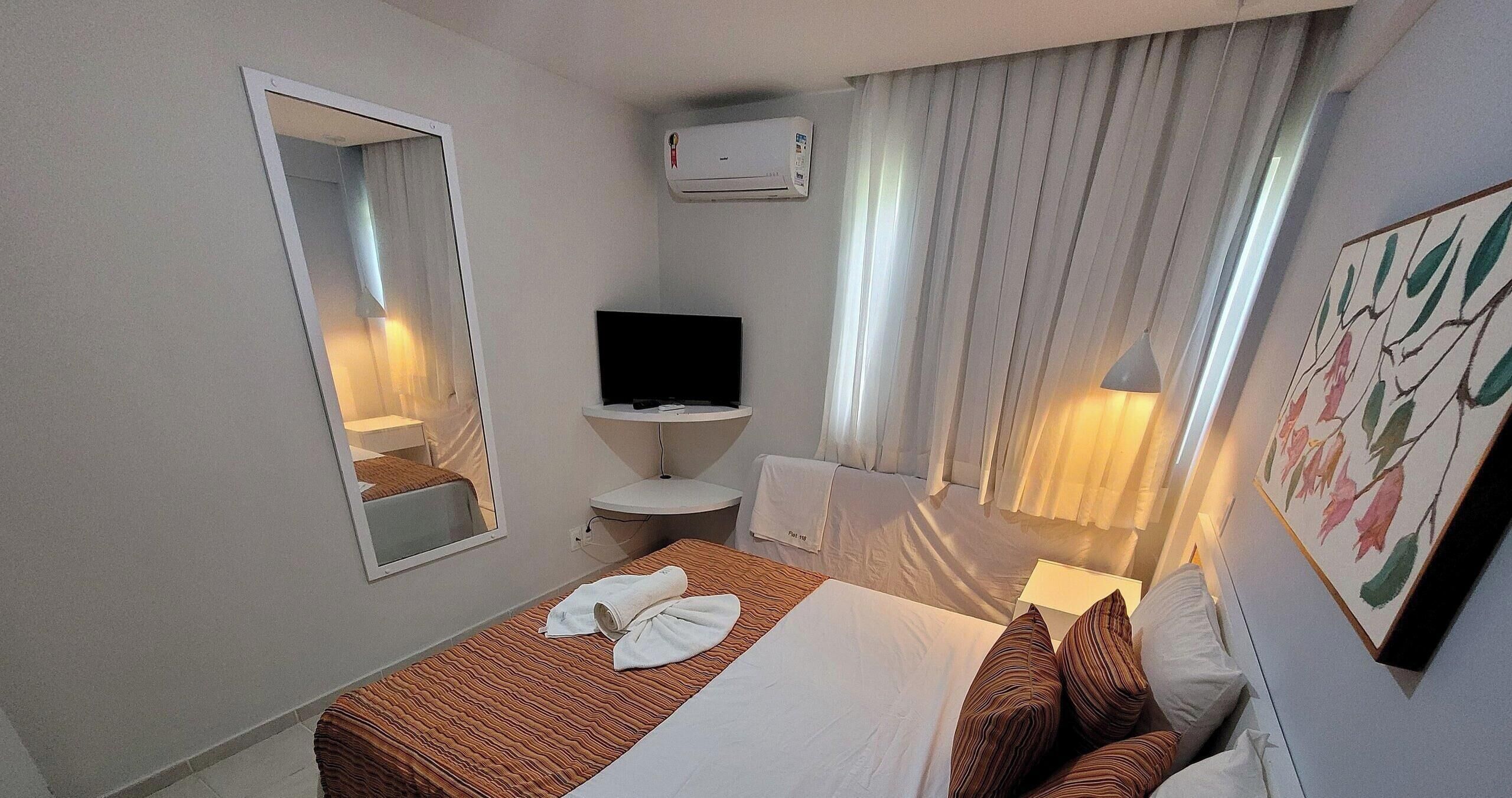 A hotel room with a bed, a TV, a mirror, a sink, and a window with curtains.