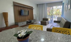 A modern living room with a flat-screen TV, a sofa, a dining table with chairs, and a balcony with outdoor furniture.