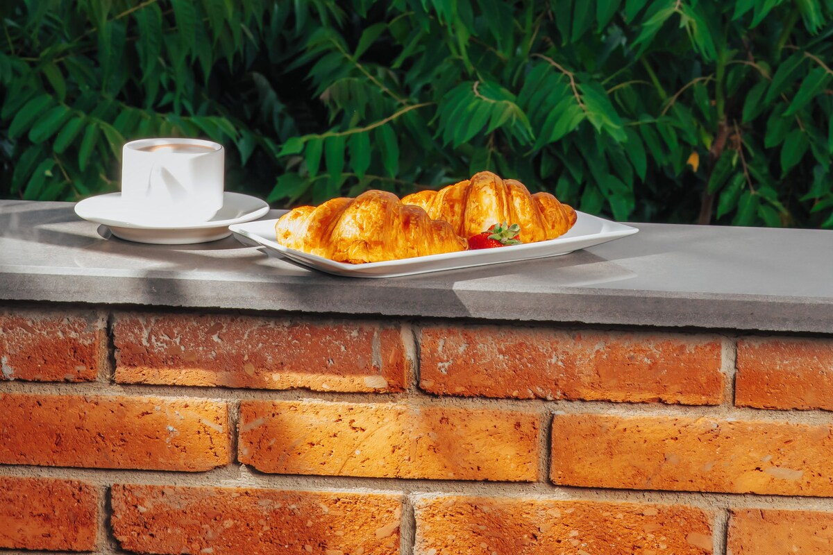 Um cenário de café da manhã com croissants e uma xícara sobre um parapeito de tijolos, com vista para uma paisagem urbana com um edifício imponente e vegetação exuberante.