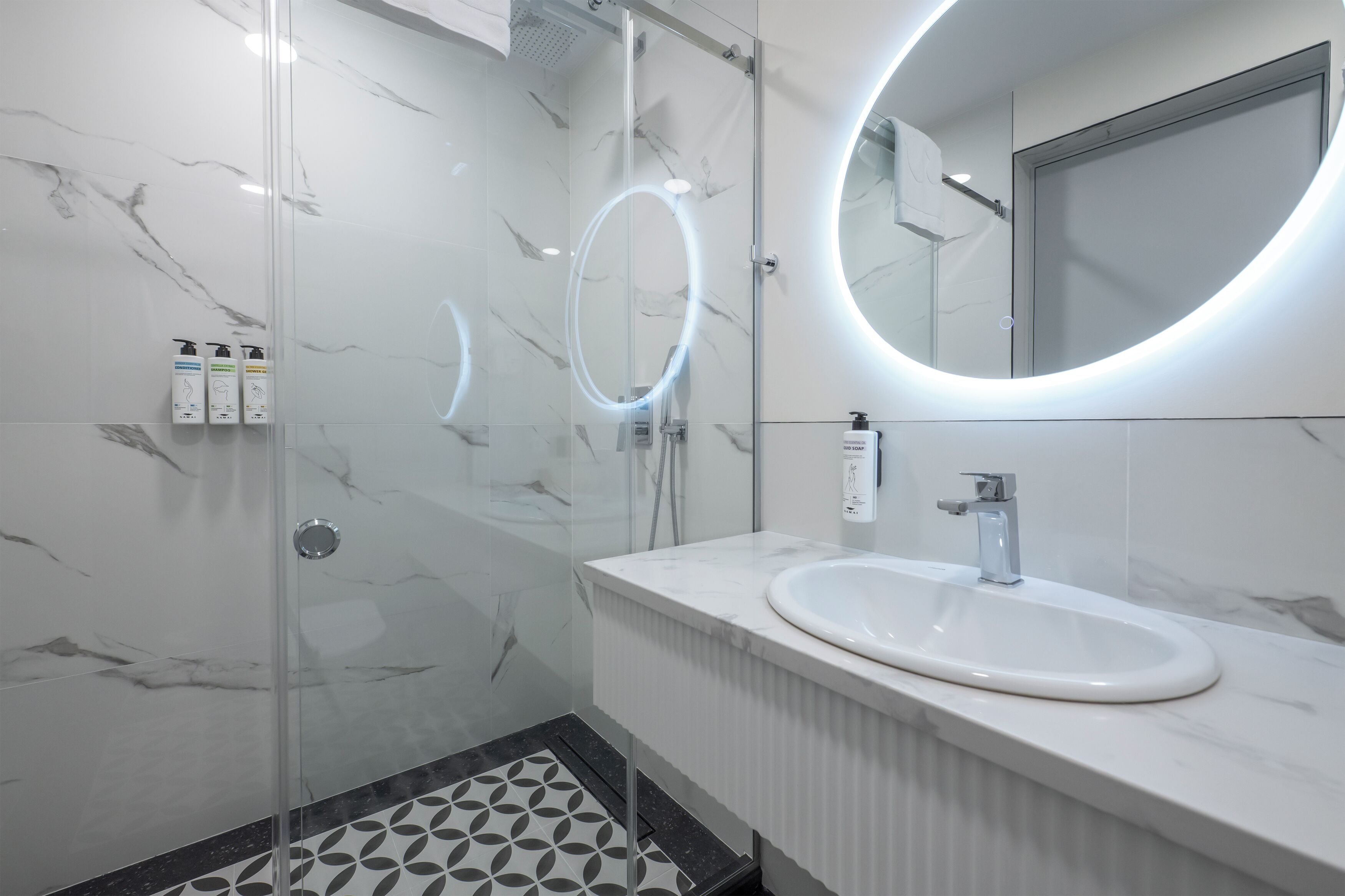 A modern bathroom with a glass-enclosed shower, a round mirror with integrated lighting, a white sink, and a marble-tiled floor.