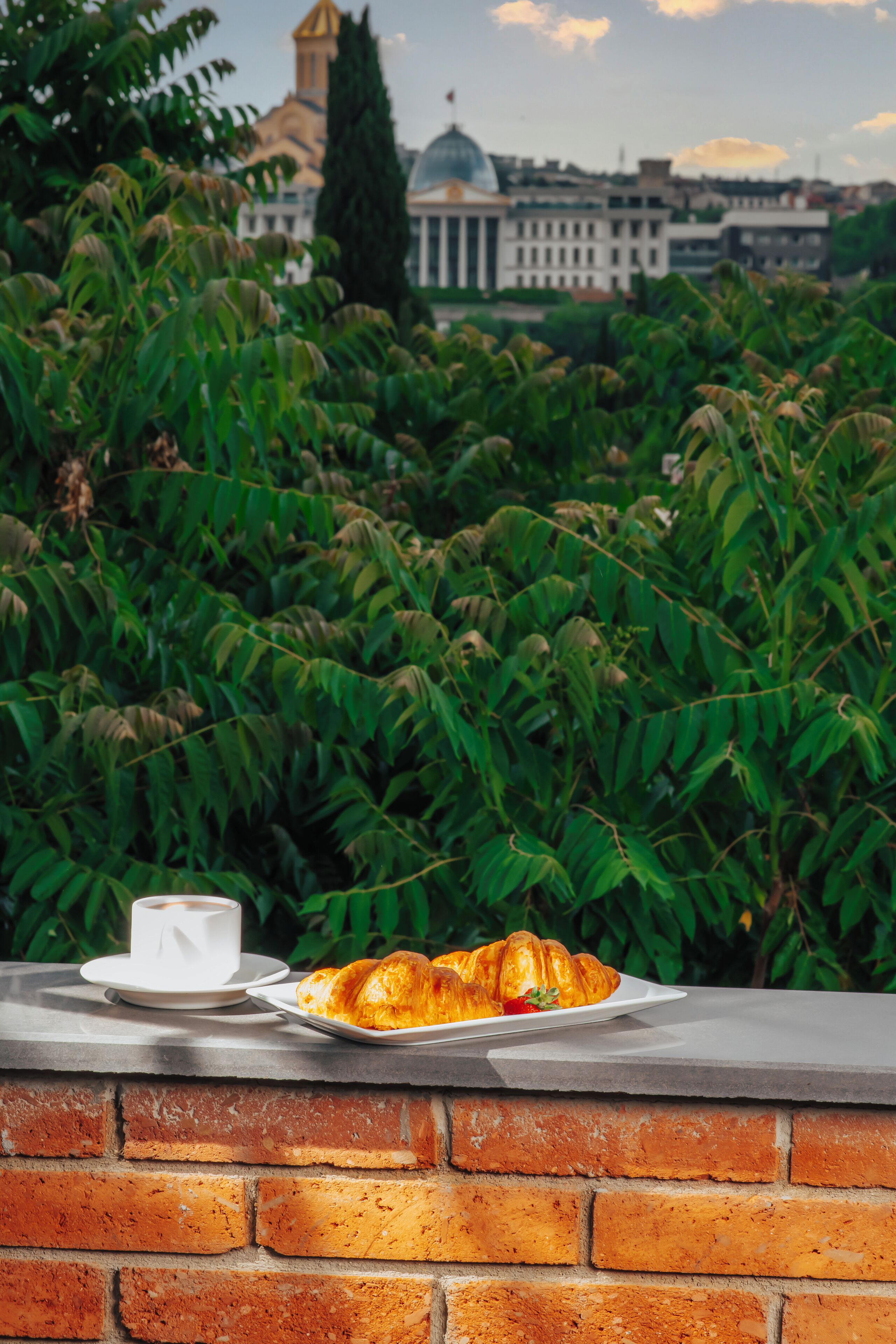 A breakfast setting with croissants and a cup on a brick ledge, overlooking a cityscape with a prominent building and lush greenery.