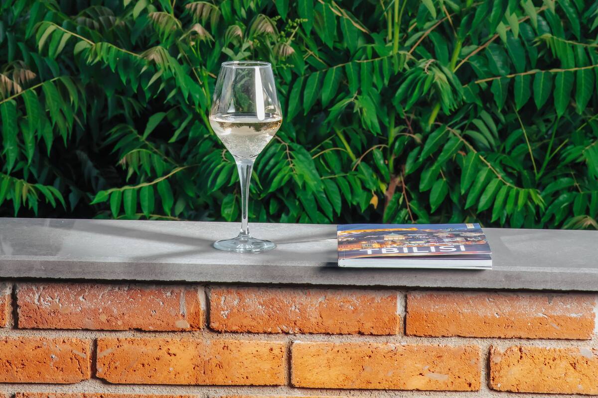 Un verre de vin blanc posé sur une étagère en briques, à côté d’un livre intitulé « Tbilissi », avec une vue sur la ville en arrière-plan.