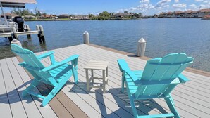 Une jetée avec des chaises Adirondack turquoise et une petite table, d’où l’on peut admirer un plan d’eau paisible, avec des bâtiments en arrière-plan.