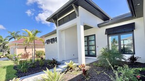 Une maison moderne à deux étages, dotée d’un toit plat, de grandes fenêtres et d’un jardin bien entretenu où poussent différentes espèces de plantes.