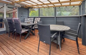 A covered outdoor seating area with a table and chairs, wooden flooring, and a view of greenery through large windows.
