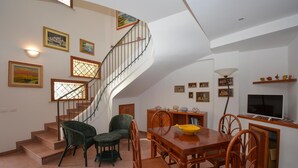 Un'ampia zona giorno con un tavolo da pranzo in legno, sedie in vimini, una scala con ringhiera e un televisore su un mobile.