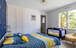 A bedroom with a bed, a crib, a chair, and a window with a view of palm trees.