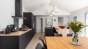 Una cocina moderna con armarios negros, una encimera de madera y una cafetera. Hay un comedor con una mesa y sillas de madera, y un jarrón con flores sobre la mesa.