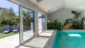 Un área de piscina interior moderna con puertas de cristal que dan a un patio trasero, un techo con tragaluces y plantas tropicales.