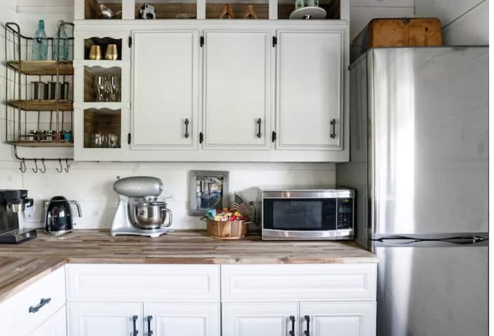 Una cocina con armarios blancos, un refrigerador de acero inoxidable, un microondas, una batidora de pie y una encimera de madera.