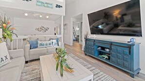 A modern living room with a large flat-screen TV, a white sofa, a blue cabinet, and a decorative plant on a coffee table. - Spacious Pet-Friendly Beach Home (Cape Canaveral)