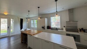 Una cocina moderna con armarios blancos, una isla de madera, luces colgantes, una campana extractora de acero inoxidable y un ventanal grande.