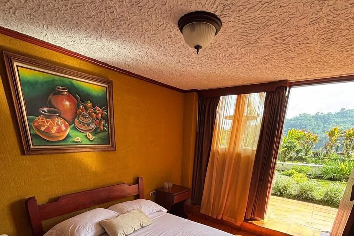 Ein Hotelzimmer mit Bett, Nachttisch, einem Gemälde und Blick auf Grünfläche.
