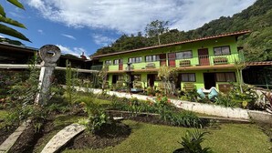 Un edificio de dos plantas con fachada verde, varios balcones y un jardín con plantas diversas y mobiliario.